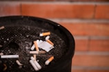A black ashtray filled with a variety of cigarette butts and ash is set against a background of red brick wall.