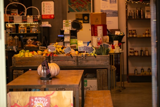 A cozy shop interior filled with fresh produce like bananas, apples, and pumpkins displayed in wooden crates. Shelves in the background hold various packaged goods and bottles. The lighting is warm, creating an inviting atmosphere.
