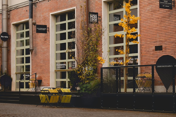 A street scene featuring a building with red brick walls and several large windows. Yellow chairs and tables are placed outside, accompanied by some potted plants. A small tree with yellow leaves adds a touch of color. Various signs are mounted on the building facade, indicating different businesses.