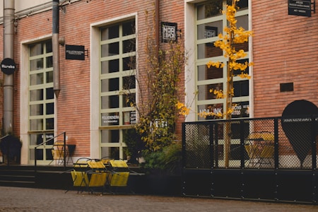 A street scene featuring a building with red brick walls and several large windows. Yellow chairs and tables are placed outside, accompanied by some potted plants. A small tree with yellow leaves adds a touch of color. Various signs are mounted on the building facade, indicating different businesses.