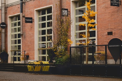 A street scene featuring a building with red brick walls and several large windows. Yellow chairs and tables are placed outside, accompanied by some potted plants. A small tree with yellow leaves adds a touch of color. Various signs are mounted on the building facade, indicating different businesses.
