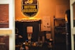 A cozy shop interior is filled with warm lighting and shelves displaying various products. The prominent sign above reads 'Bottega Biologica Organic Market' in yellow against a black background. Below the sign, there are informational posters on the wall, and shelves stocked with items like bottles and boxes. A variety of products, including fruits, are visible on a counter.