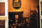 A cozy shop interior is filled with warm lighting and shelves displaying various products. The prominent sign above reads 'Bottega Biologica Organic Market' in yellow against a black background. Below the sign, there are informational posters on the wall, and shelves stocked with items like bottles and boxes. A variety of products, including fruits, are visible on a counter.