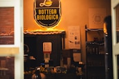 A cozy shop interior is filled with warm lighting and shelves displaying various products. The prominent sign above reads 'Bottega Biologica Organic Market' in yellow against a black background. Below the sign, there are informational posters on the wall, and shelves stocked with items like bottles and boxes. A variety of products, including fruits, are visible on a counter.