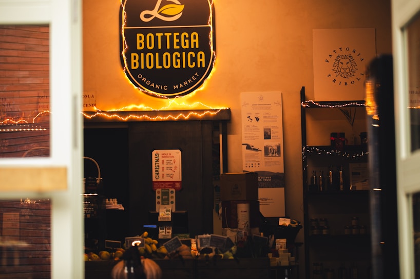 A cozy shop interior is filled with warm lighting and shelves displaying various products. The prominent sign above reads 'Bottega Biologica Organic Market' in yellow against a black background. Below the sign, there are informational posters on the wall, and shelves stocked with items like bottles and boxes. A variety of products, including fruits, are visible on a counter.