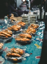 A market stall displaying several plastic containers filled with cooked prawns. The containers are arranged on a colorful tablecloth with graphic designs and text. In the background, there are other food items and packaging visible.