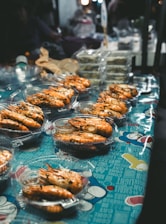 A market stall displaying several plastic containers filled with cooked prawns. The containers are arranged on a colorful tablecloth with graphic designs and text. In the background, there are other food items and packaging visible.
