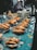 A market stall displaying several plastic containers filled with cooked prawns. The containers are arranged on a colorful tablecloth with graphic designs and text. In the background, there are other food items and packaging visible.