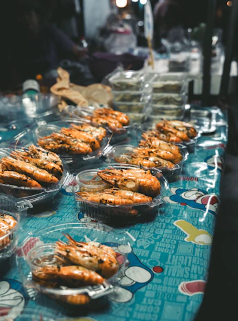 A market stall displaying several plastic containers filled with cooked prawns. The containers are arranged on a colorful tablecloth with graphic designs and text. In the background, there are other food items and packaging visible.