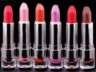 A row of six lipsticks in varying shades displayed against a black background. The lipsticks are housed in metallic silver tubes, with colors including red, pink, orange, and brown. Each lipstick has a distinct color, creating a gradient effect.