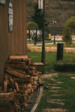 A stack of firewood is neatly arranged against the side of a wooden building. A pathway of stones leads into the grass beyond, with a lamppost standing nearby. The scene includes various plants and a few signs in the background, suggesting a park or garden setting.
