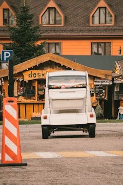 A small white vehicle is parked in front of a wooden kiosk with the word 'kiosk' written on it. The background features a brown building with triangular windows, and there is a large pine tree on the left side. An orange and white striped traffic cone is visible in the foreground.