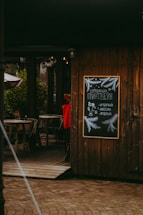 A charming outdoor cafe setting with wooden walls and decor. It features a chalkboard menu with festive illustrations and the text written in Russian. The entrance has wicker chairs and tables, creating a cozy and inviting atmosphere.