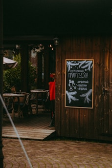 A charming outdoor cafe setting with wooden walls and decor. It features a chalkboard menu with festive illustrations and the text written in Russian. The entrance has wicker chairs and tables, creating a cozy and inviting atmosphere.