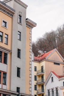 A collection of residential buildings with varied architectural styles, featuring neutral tones such as beige, cream, and light yellow. The buildings have multiple windows and some balconies. Located in front of a background of bare trees, indicating a winter or early spring setting.