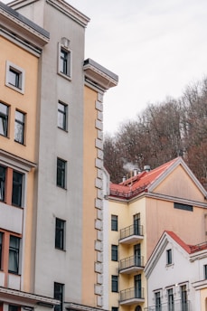 A collection of residential buildings with varied architectural styles, featuring neutral tones such as beige, cream, and light yellow. The buildings have multiple windows and some balconies. Located in front of a background of bare trees, indicating a winter or early spring setting.