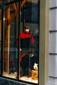 A storefront display features a mannequin dressed in a red and black ski outfit, complete with goggles and holding a snowboard. The scene is set against a snowy background with decorative logs and artificial snow, creating a winter sports theme.