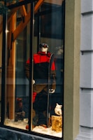 A storefront display features a mannequin dressed in a red and black ski outfit, complete with goggles and holding a snowboard. The scene is set against a snowy background with decorative logs and artificial snow, creating a winter sports theme.