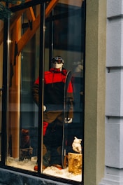 A storefront display features a mannequin dressed in a red and black ski outfit, complete with goggles and holding a snowboard. The scene is set against a snowy background with decorative logs and artificial snow, creating a winter sports theme.