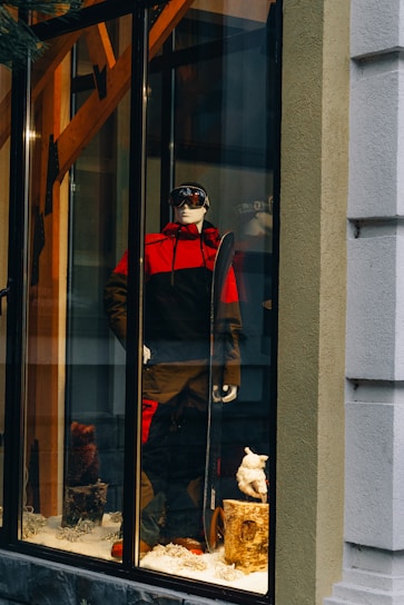 A storefront display features a mannequin dressed in a red and black ski outfit, complete with goggles and holding a snowboard. The scene is set against a snowy background with decorative logs and artificial snow, creating a winter sports theme.