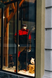 A storefront display features a mannequin dressed in a red and black ski outfit, complete with goggles and holding a snowboard. The scene is set against a snowy background with decorative logs and artificial snow, creating a winter sports theme.