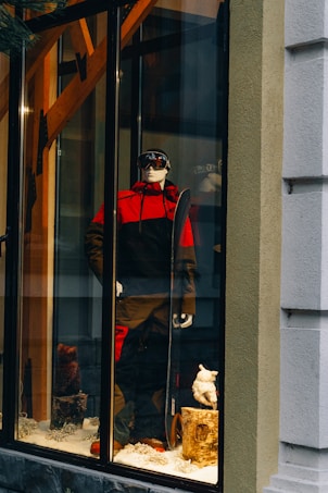 A storefront display features a mannequin dressed in a red and black ski outfit, complete with goggles and holding a snowboard. The scene is set against a snowy background with decorative logs and artificial snow, creating a winter sports theme.