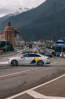A busy street scene with cars traveling in both directions in a mountainous area. A white car labeled 'Yandex Go' is prominently visible in the foreground, possibly at a turn or intersection. Buildings with architectural features and signs line the road, and snow-capped mountains can be seen in the background under overcast skies.