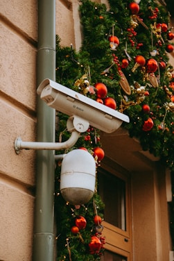 A security camera is mounted on an exterior wall with festive holiday decorations. Green garlands adorned with red and gold ornaments and small bird decorations hang nearby, enhancing the holiday atmosphere.