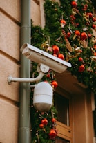 A security camera is mounted on an exterior wall with festive holiday decorations. Green garlands adorned with red and gold ornaments and small bird decorations hang nearby, enhancing the holiday atmosphere.