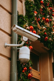 A security camera is mounted on an exterior wall with festive holiday decorations. Green garlands adorned with red and gold ornaments and small bird decorations hang nearby, enhancing the holiday atmosphere.