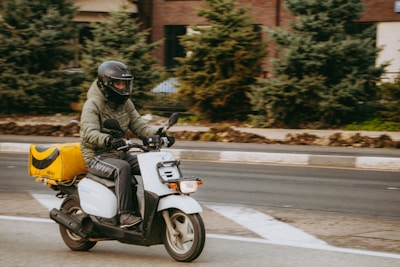 A person wearing a helmet and jacket rides a white scooter on a road. They carry a yellow delivery box attached to the back. The background features greenery and a building.