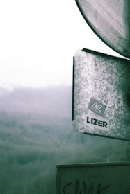 A metal sign with a weathered texture featuring stickers, one reading 'LIZER' and another with a geometric design and text 'ART IN CLO'. The background is misty, suggesting a foggy landscape or mountainous area.