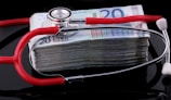 A stethoscope with red tubing placed over a stack of 20-euro banknotes, symbolizing a connection between healthcare and finances.