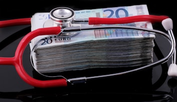 A stethoscope with red tubing placed over a stack of 20-euro banknotes, symbolizing a connection between healthcare and finances.