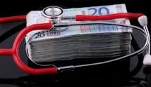 A stethoscope with red tubing placed over a stack of 20-euro banknotes, symbolizing a connection between healthcare and finances.