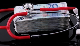 A stethoscope with red tubing placed over a stack of 20-euro banknotes, symbolizing a connection between healthcare and finances.