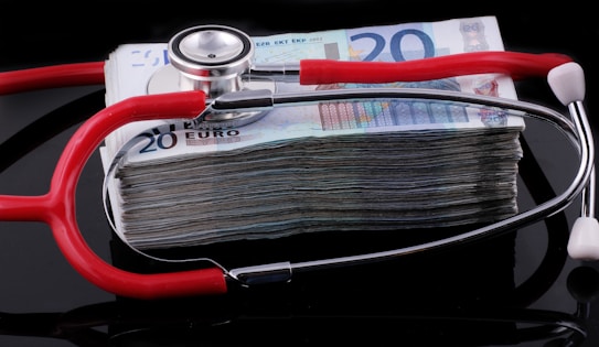 A stethoscope with red tubing placed over a stack of 20-euro banknotes, symbolizing a connection between healthcare and finances.