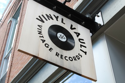 A wooden sign with a retro design hangs outside a building, featuring the words 'Vinyl Vault Vintage Records' surrounding an image of a vinyl record. The sign is affixed to a metal bracket mounted on the exterior wall, which is made of red brick with a glass window nearby.