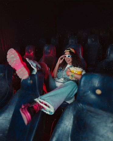 A person is reclining in a dimly lit theater seat, casually dressed in a denim jacket and 3D glasses, with feet up on another chair. They are holding a bucket of popcorn and appear to be enjoying themselves, with a playful expression.