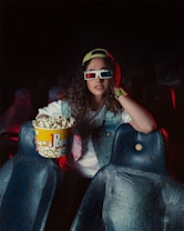 A person wearing 3D glasses and a denim vest holds a large tub of popcorn in a dimly lit cinema. The individual is also wearing a watch and a cap turned backward, exuding a casual and relaxed vibe. The seats around them are empty, and there is a slight red and blue lighting effect.