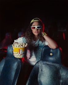 A person wearing 3D glasses and a denim vest holds a large tub of popcorn in a dimly lit cinema. The individual is also wearing a watch and a cap turned backward, exuding a casual and relaxed vibe. The seats around them are empty, and there is a slight red and blue lighting effect.