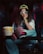 A person with curly hair wears 3D glasses and a yellow cap sitting in a theater seat, holding a large tub of popcorn. Dressed in casual denim and a white T-shirt, one leg is propped up on another seat, revealing bright pink shoes. The setting is dimly lit, suggesting a cinema environment.