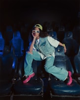 A person sitting in a dimly lit theatre on worn-out seats, wearing a denim jacket and jeans. The individual sports bright pink sneakers, a yellow cap, and 3D glasses, displaying a casual and relaxed posture.