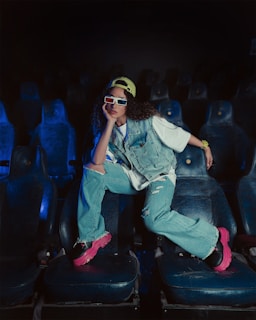 A person sitting in a dimly lit theatre on worn-out seats, wearing a denim jacket and jeans. The individual sports bright pink sneakers, a yellow cap, and 3D glasses, displaying a casual and relaxed posture.