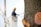 A close-up of a tree trunk with visible cut branches on the right side. The wood is exposed, showing circular cuts with sap oozing out. In the background, there is a worker wearing a yellow hard hat, blurred and standing in an elevated bucket, possibly trimming or inspecting the tree.