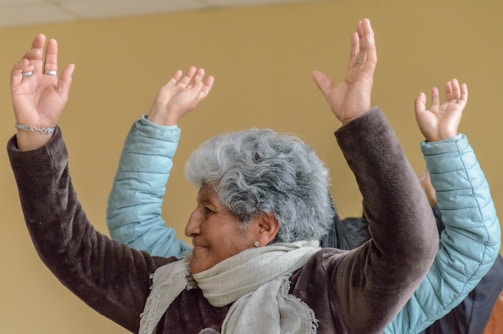 An elderly person, wearing a brown jacket and a scarf, lifts their arms in what seems to be a joyful or celebratory gesture. Several others in the background also have their arms raised. The environment is indoors with a neutral-colored wall.