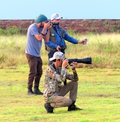 Three photographers are outdoors on a grassy field. They are dressed in casual and camouflage clothing, and each holds a camera with a long lens. One photographer is kneeling while the other two are standing. The background shows a mix of grass and a blurry horizon.