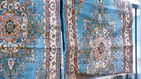 Two intricately designed carpets hang side by side, featuring a rich pattern of floral and geometric motifs. The carpets display a vivid mix of colors, with predominant hues of blue, orange, and white. The texture appears plush and detailed, suggesting a high level of craftsmanship.