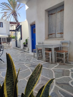 A quaint and quiet outdoor setting with a small wooden table and chairs placed against a whitewashed building. The stone pathway leads past a blue door and shuttered window, surrounded by greenery and a tall palm tree. Nearby buildings feature classic Mediterranean architecture.