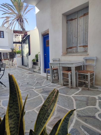 A sunlit Mediterranean terrace with pastel-colored walls, a table set with Le Coquers beverages, and olive branches swaying gently in the breeze.